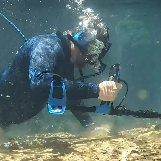 Diver underwater holding a smartphone with a Garrett metal detector attached.